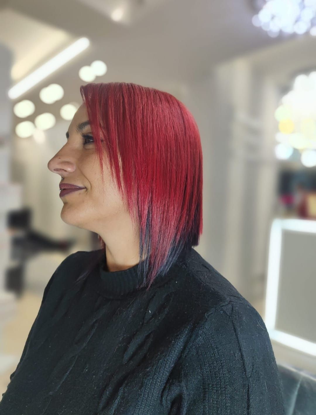 Ritratto di profilo di una persona con i capelli rosso acceso, seduta su una poltrona da parrucchiere e con indosso un mantello nero.