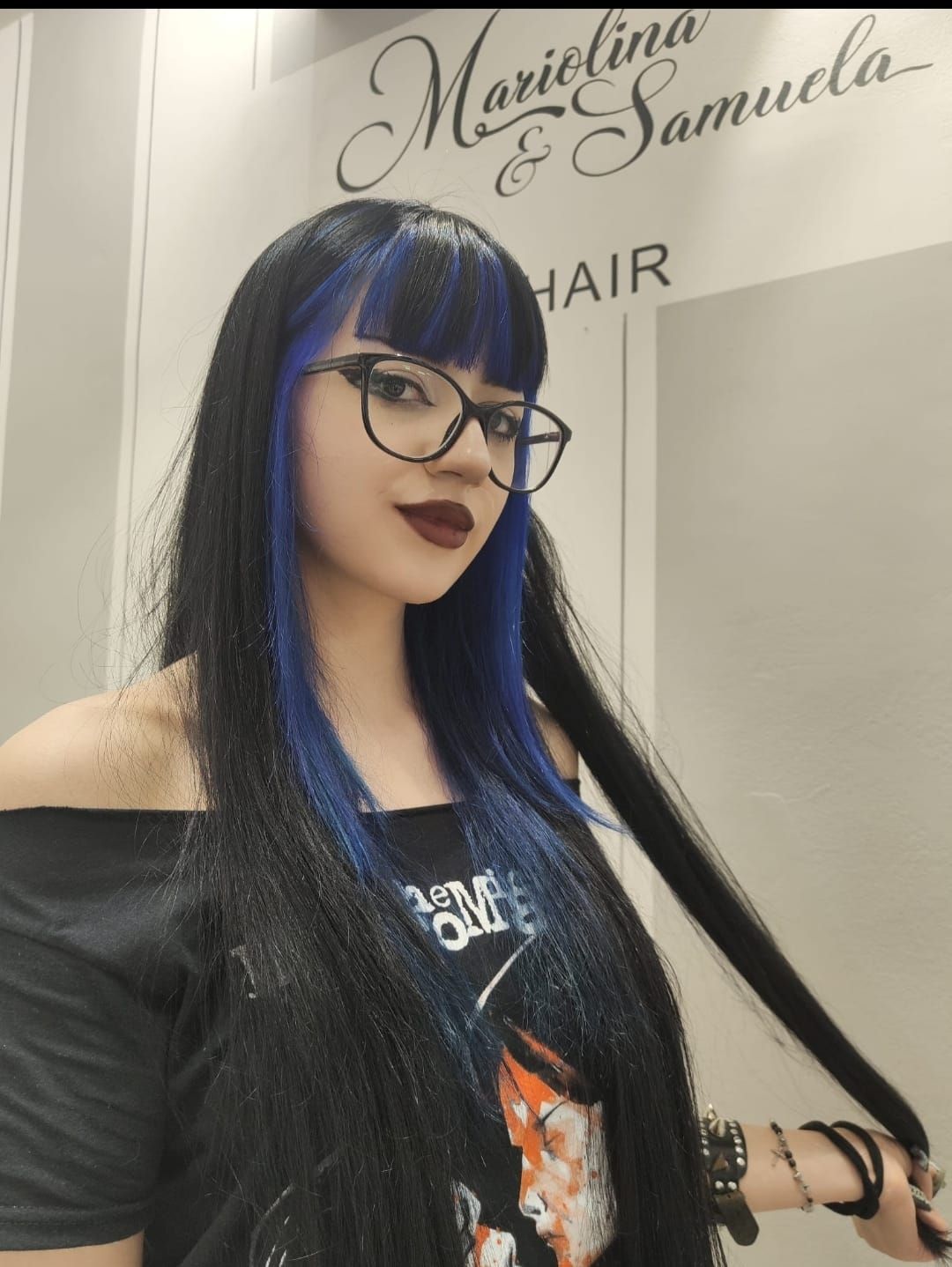 Donna con lunghi capelli neri e blu e occhiali in posa su una sedia da parrucchiere