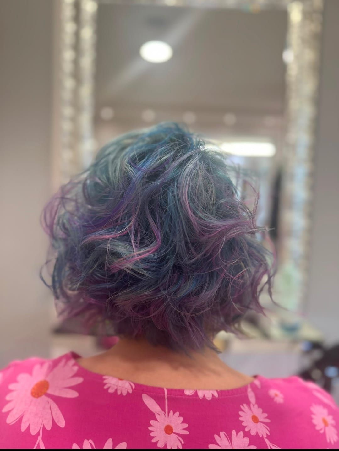 Persona con capelli corti blu e viola, vista di spalle, che indossa una maglietta rosa a fiori davanti a uno specchio.