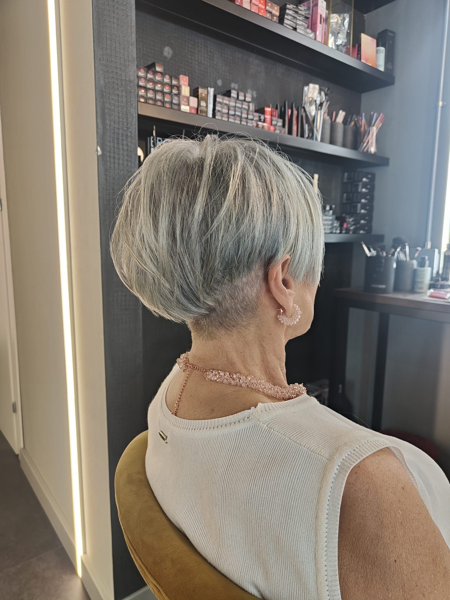 Persona con capelli corti e argentati seduta su una poltrona da parrucchiere, vista di spalle, con prodotti sugli scaffali nelle vicinanze.