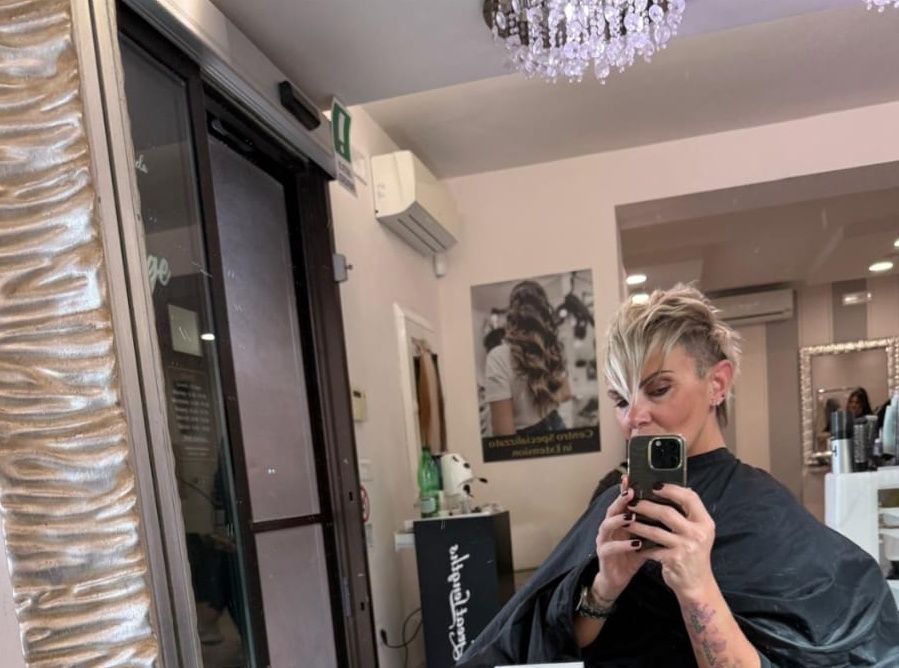 Una persona si scatta un selfie allo specchio seduta su una poltrona da parrucchiere, con i capelli biondi acconciati e un mantello scuro.