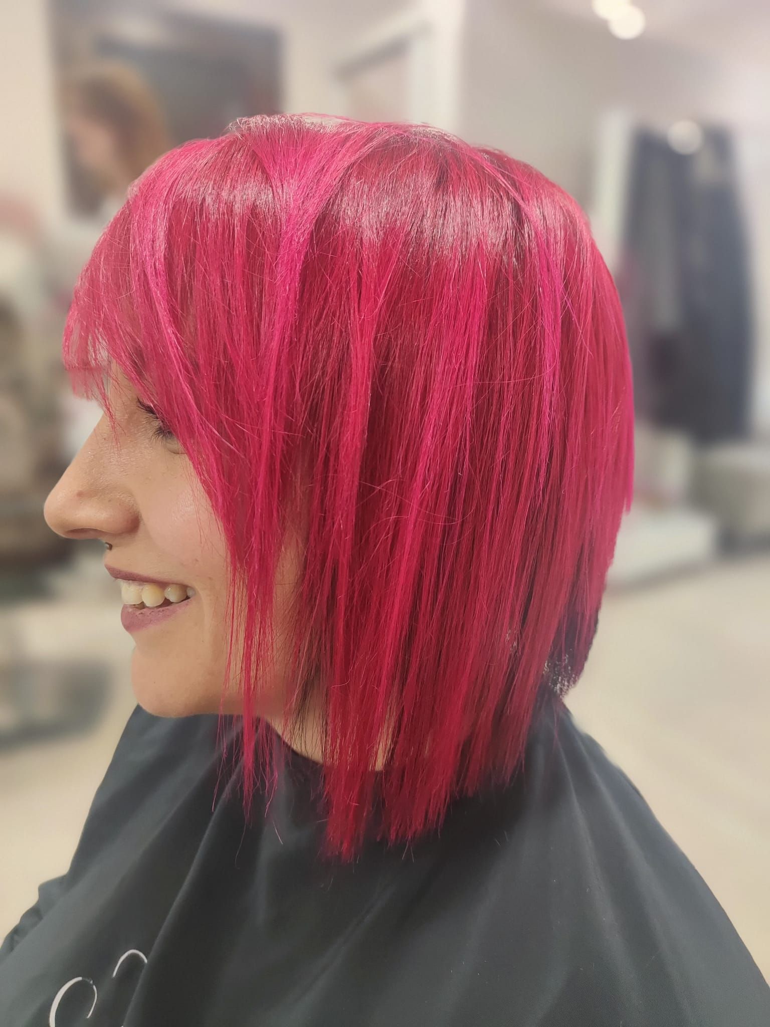 Profilo laterale di una persona con i capelli rosa acceso e un taglio a caschetto corto, seduta su una poltrona da parrucchiere, sorridente.
