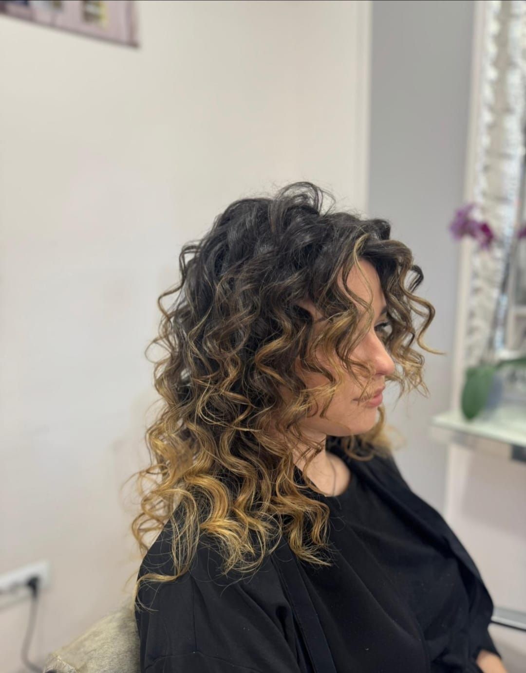 Vista di profilo di una persona con capelli scuri ricci e mèches bionde, seduta su una poltrona da parrucchiere.