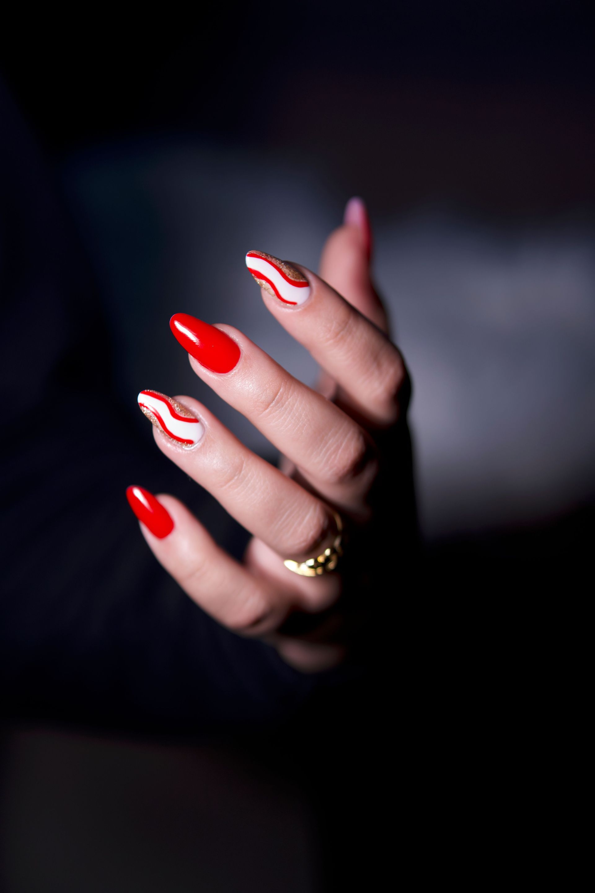 Mano con lunghe unghie rosse e bianche su sfondo scuro, con un anello d'oro al dito.