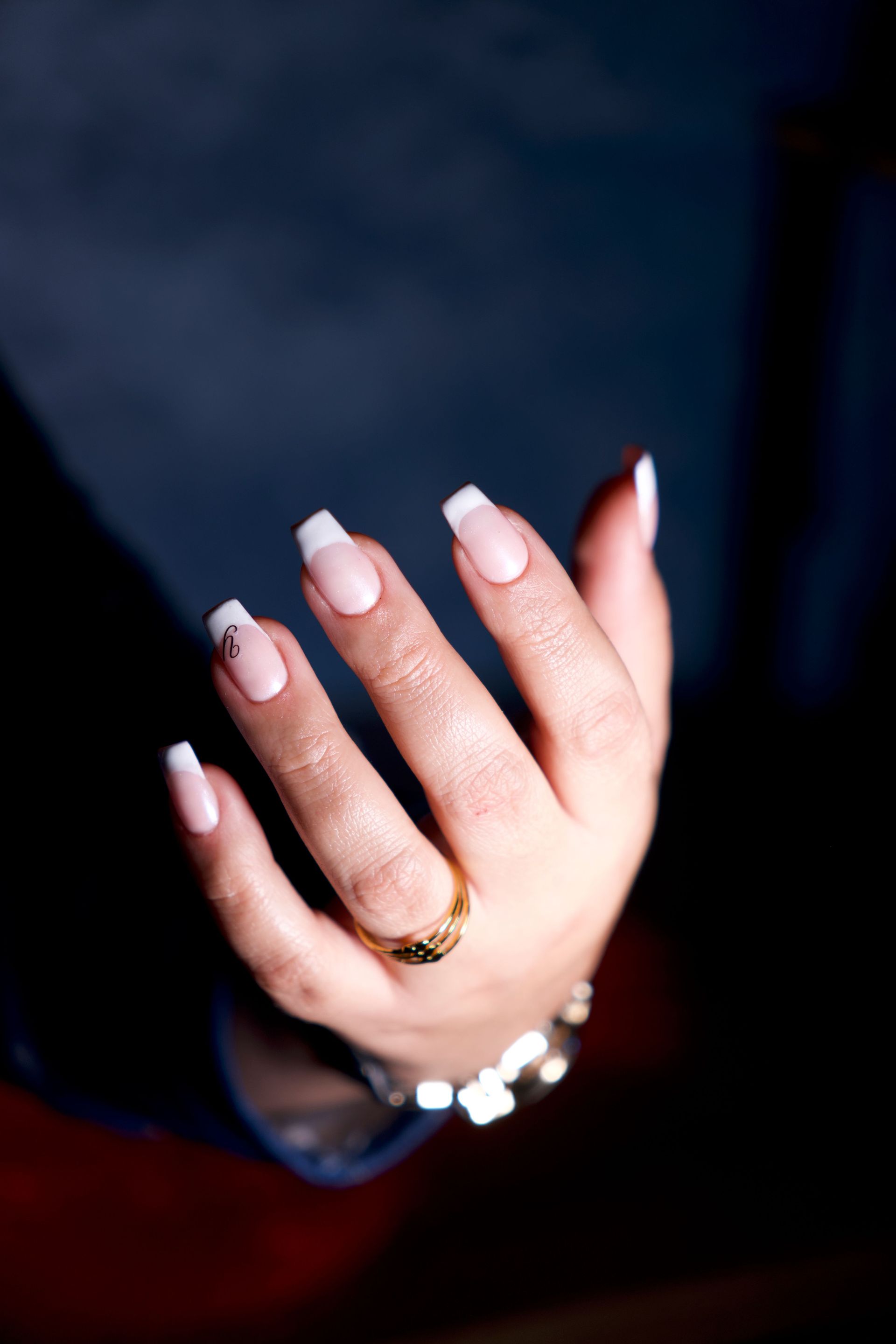 Primo piano di una mano con unghie bianche curate e un anello d'oro su sfondo scuro.