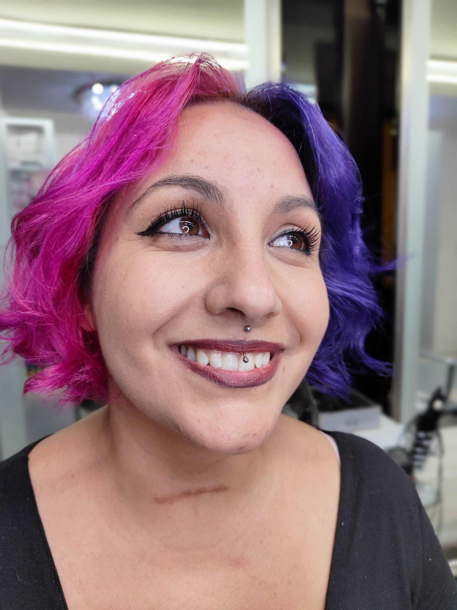Persona sorridente in un ambiente interno con capelli metà rosa e metà viola e rossetto scuro.