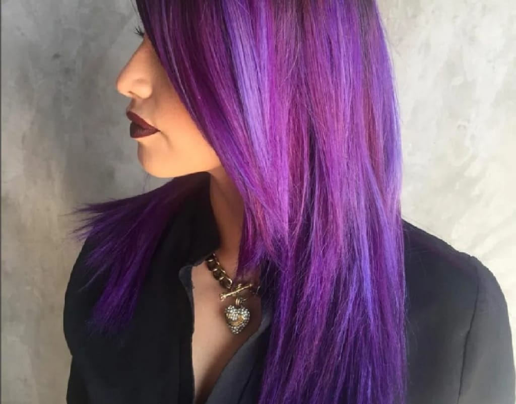 Vista di profilo di una persona con lunghi capelli viola, rossetto scuro e giacca nera su sfondo grigio.