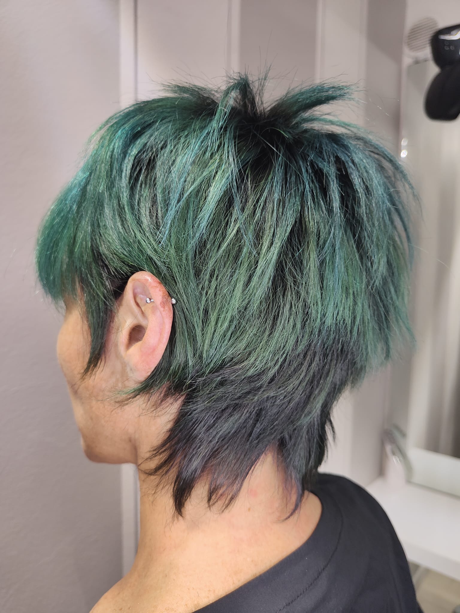 Vista laterale di una persona con capelli corti, scalati, verdi e neri, riflessa in uno specchio di un salone di bellezza.