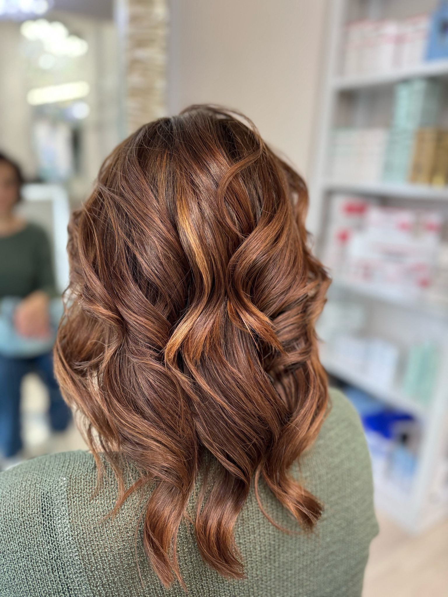 Capelli castani ondulati con riflessi caramello in un salone, visti da dietro.