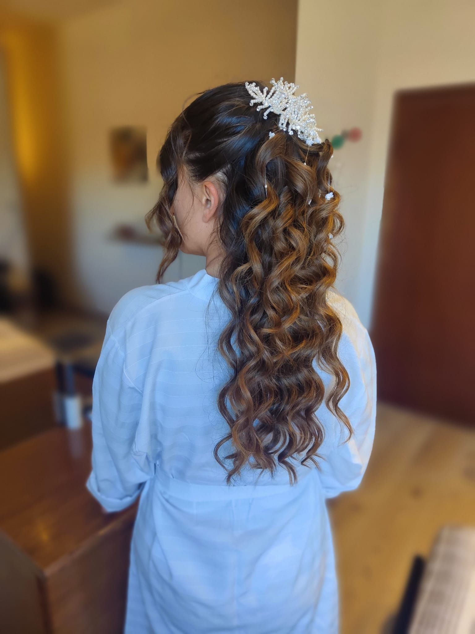 Vista posteriore di una persona con un abito azzurro chiaro e capelli ricci, in piedi in un ambiente interno con un accessorio floreale tra i capelli.