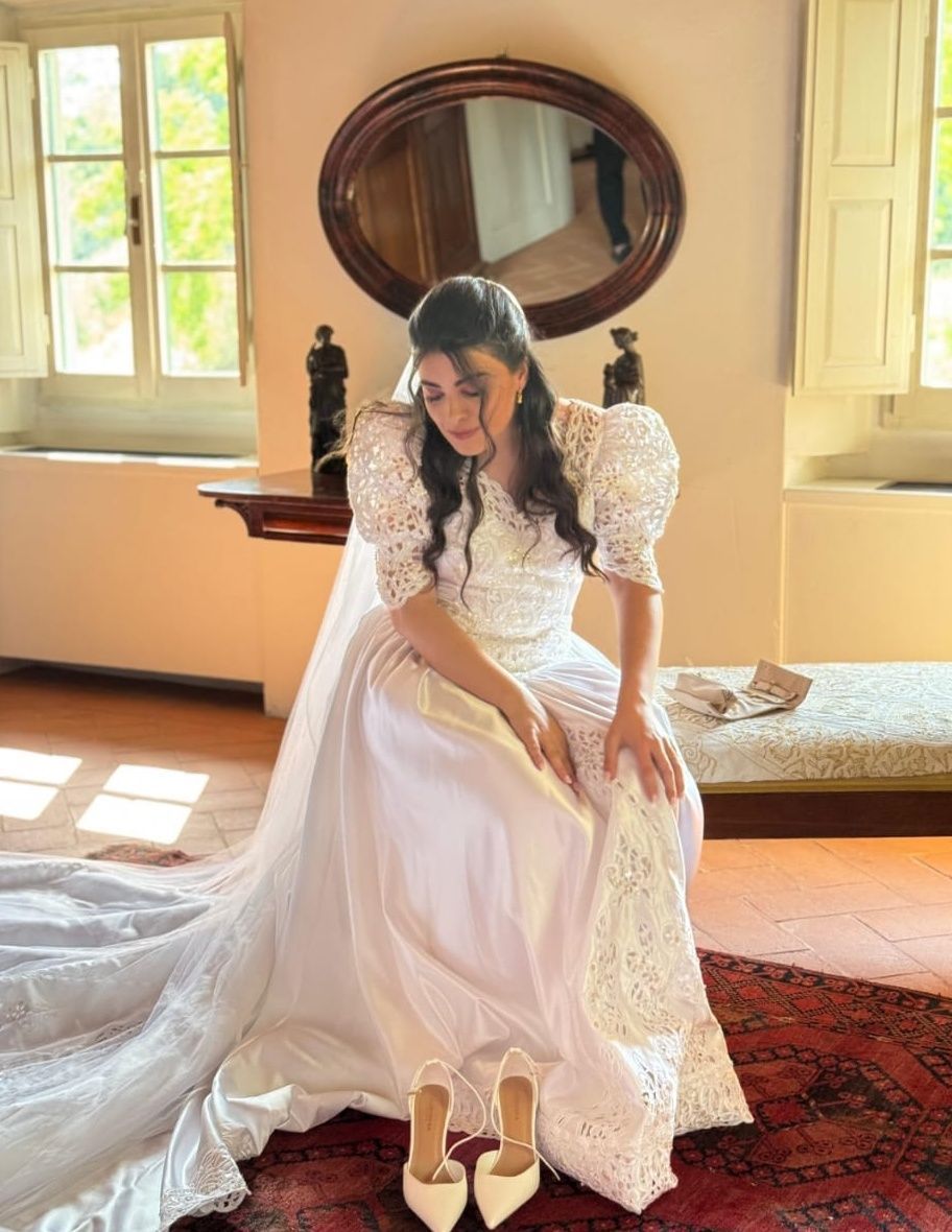 Donna in abito di pizzo bianco seduta su un letto in una camera da letto vintage illuminata dal sole, scarpe sul pavimento lì vicino.