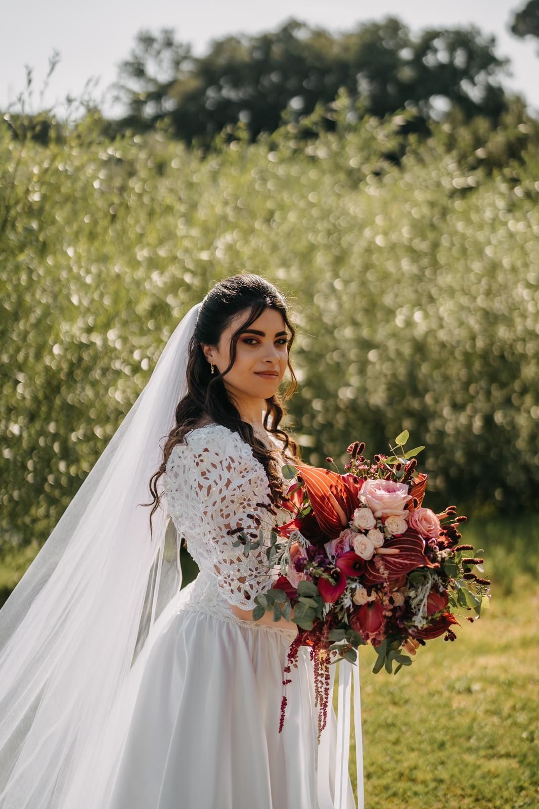 Sposa in abito di pizzo bianco che tiene in mano un bouquet di fiori rossi e rosa all'aperto in un prato.