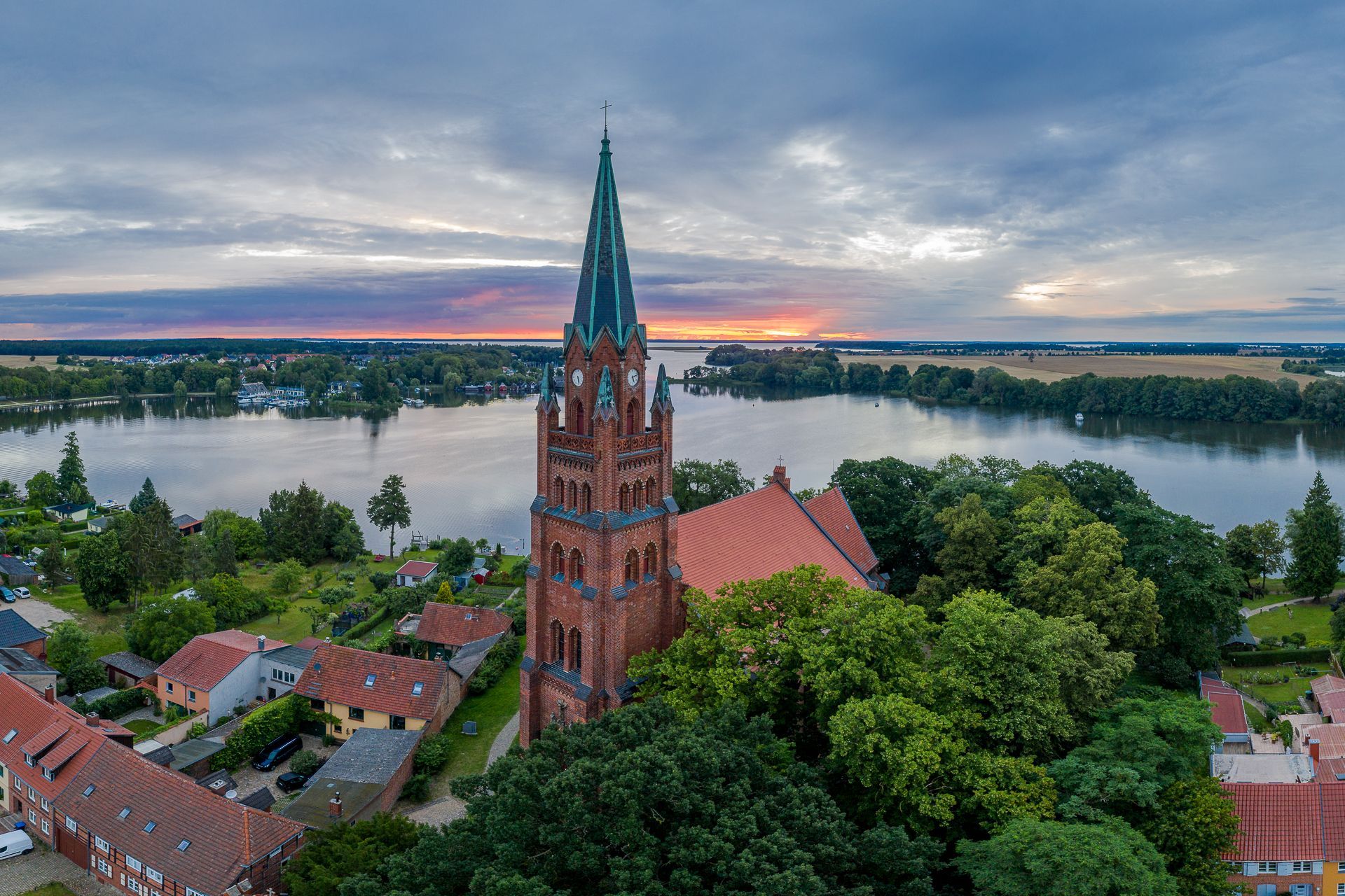 Eine Backsteinkirche mit hohem Turm, rotem Dach und grünen Bäumen, neben einem See bei Sonnenuntergang.