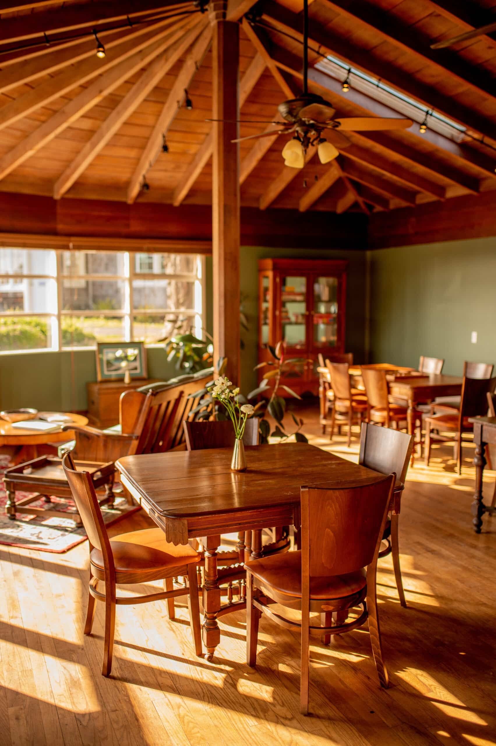 Wooden dining room with tables, chairs, and a central pillar. Sunlight streams through a window.