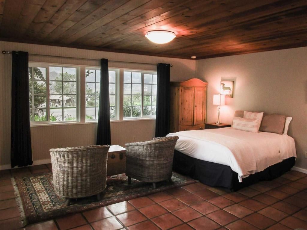 Bedroom with a bed, two wicker chairs, a rug, large windows, wood ceiling, and terracotta floor.