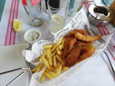 Fish and chips served in a wire basket, with a lemon, tartar sauce, and a drink on a table.