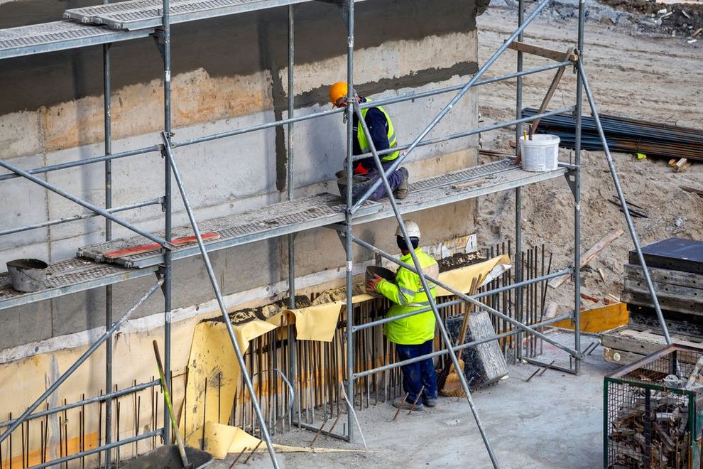Construction workers on scaffolding repairing a concrete wall, one in a yellow safety vest.