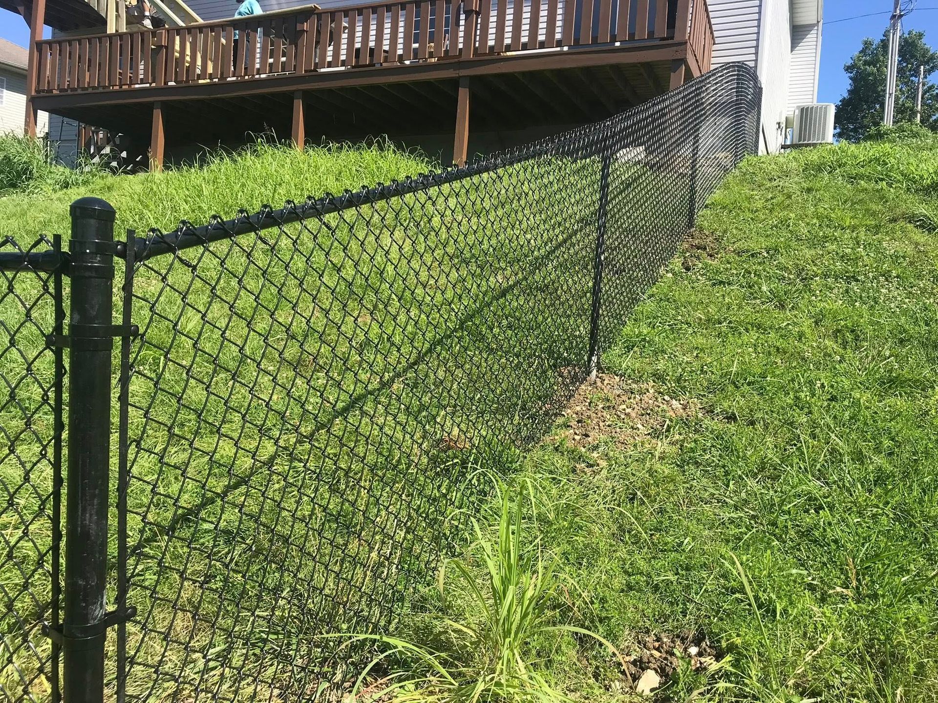 A black chain-link fence runs up a grassy hill toward a wooden deck attached to a house.