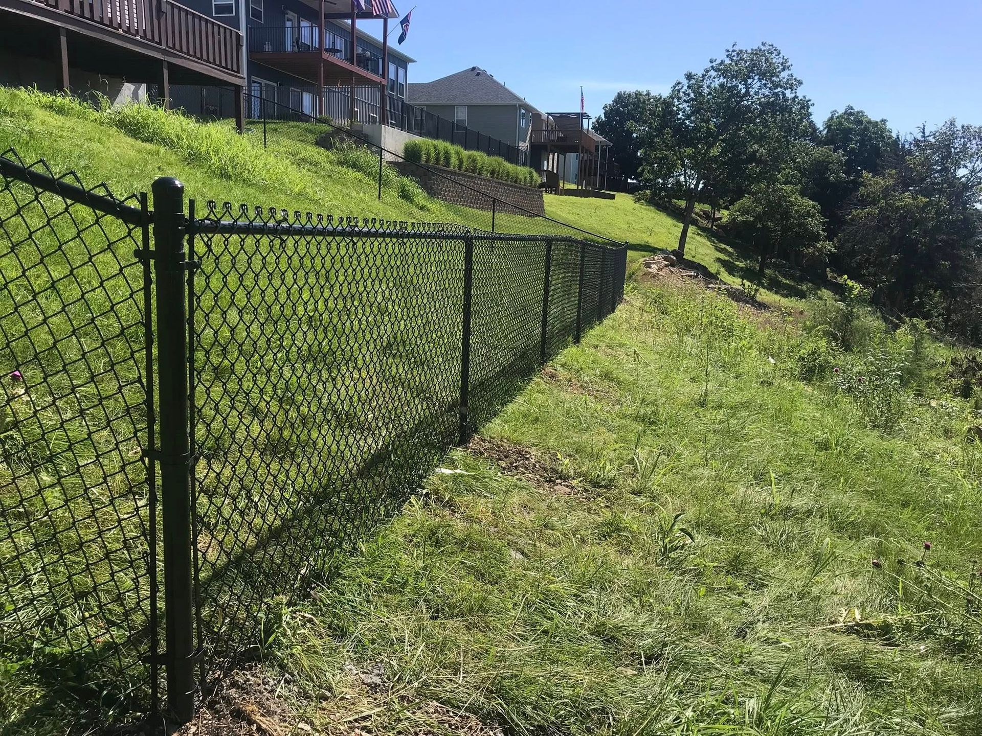 A black chain-link fence runs along a grassy, sloped hill toward houses under a clear blue sky.