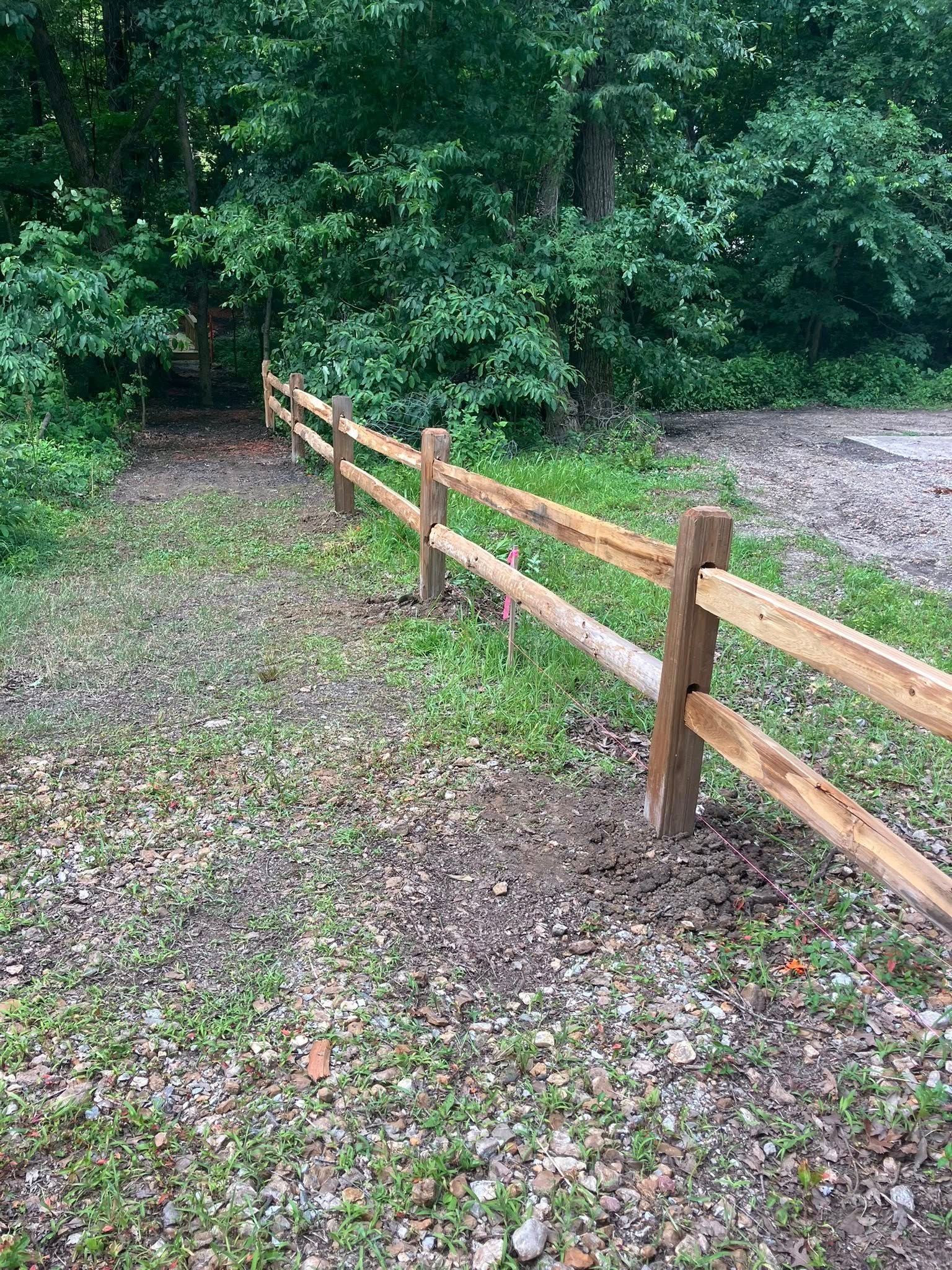 A wooden two-rail post fence lines a path leading toward a shaded forest edge.