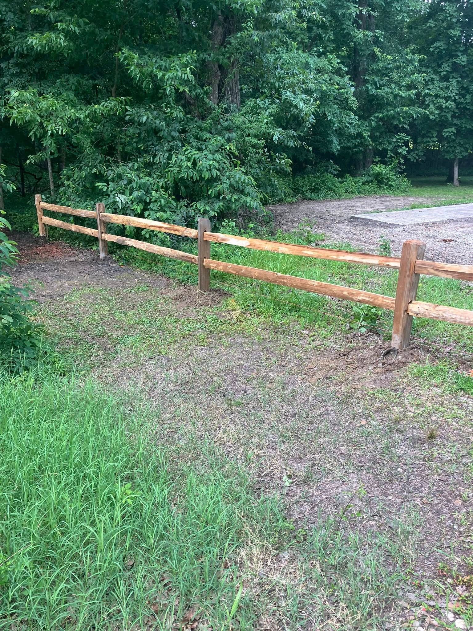 A wooden split-rail fence runs through a grassy, gravel-lined area in front of a line of trees.