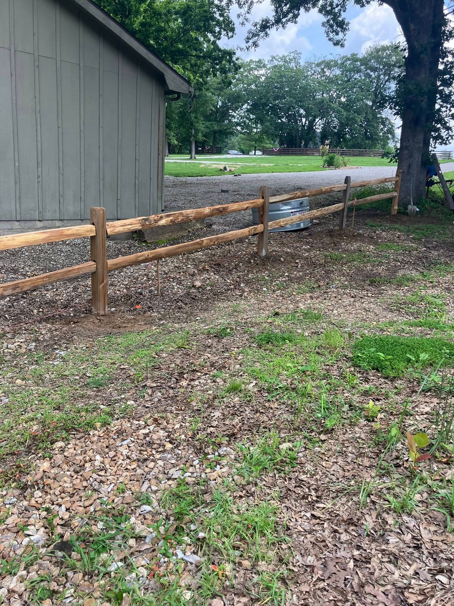 A two-rail wooden fence runs past the side of a gray shed, with a large tree and a grassy yard in the background.