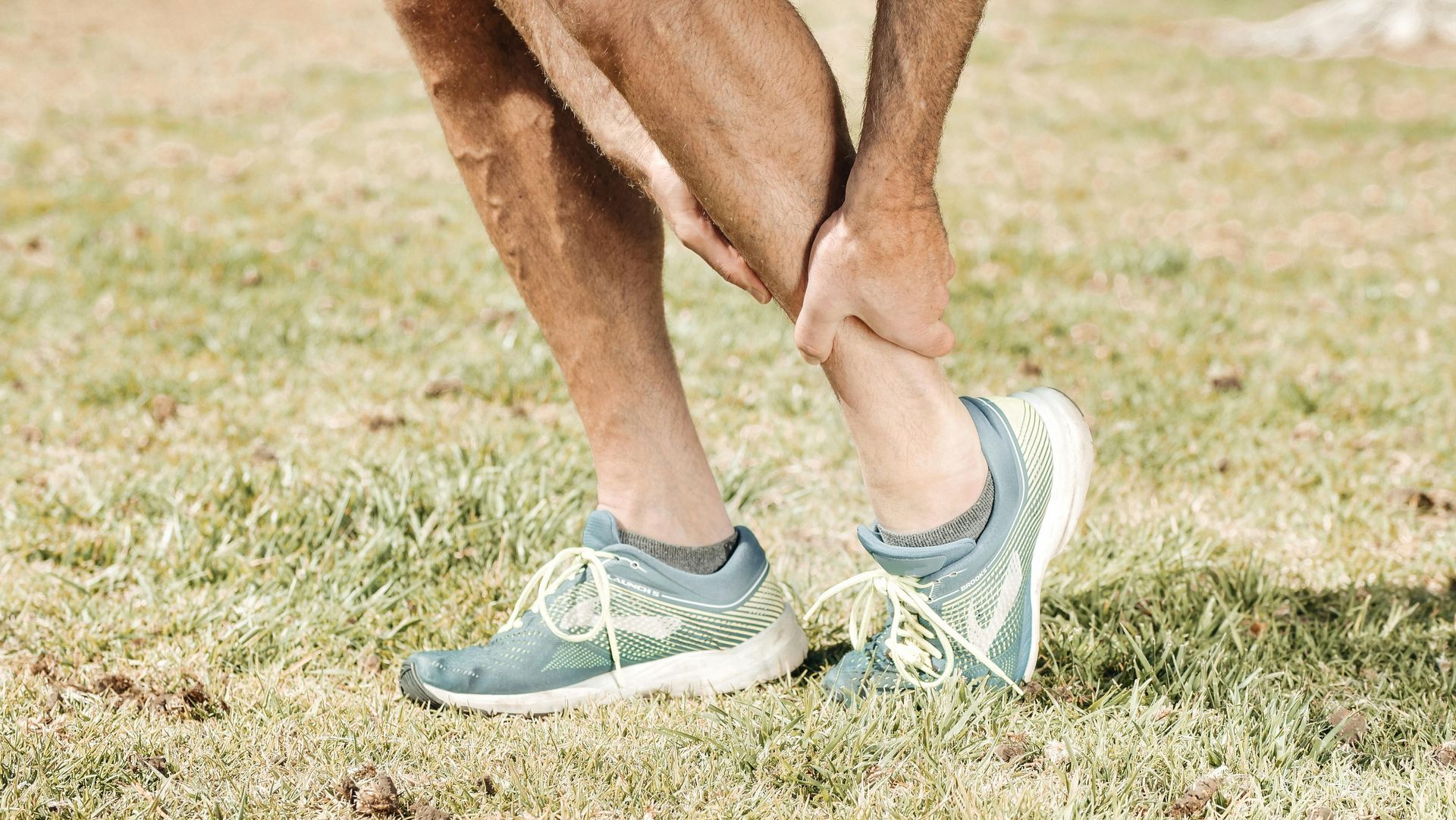 A man is holding his ankle in pain while standing in the grass — Newcastle Foot & Ankle Clinic Charlestown In Belmont, NSW