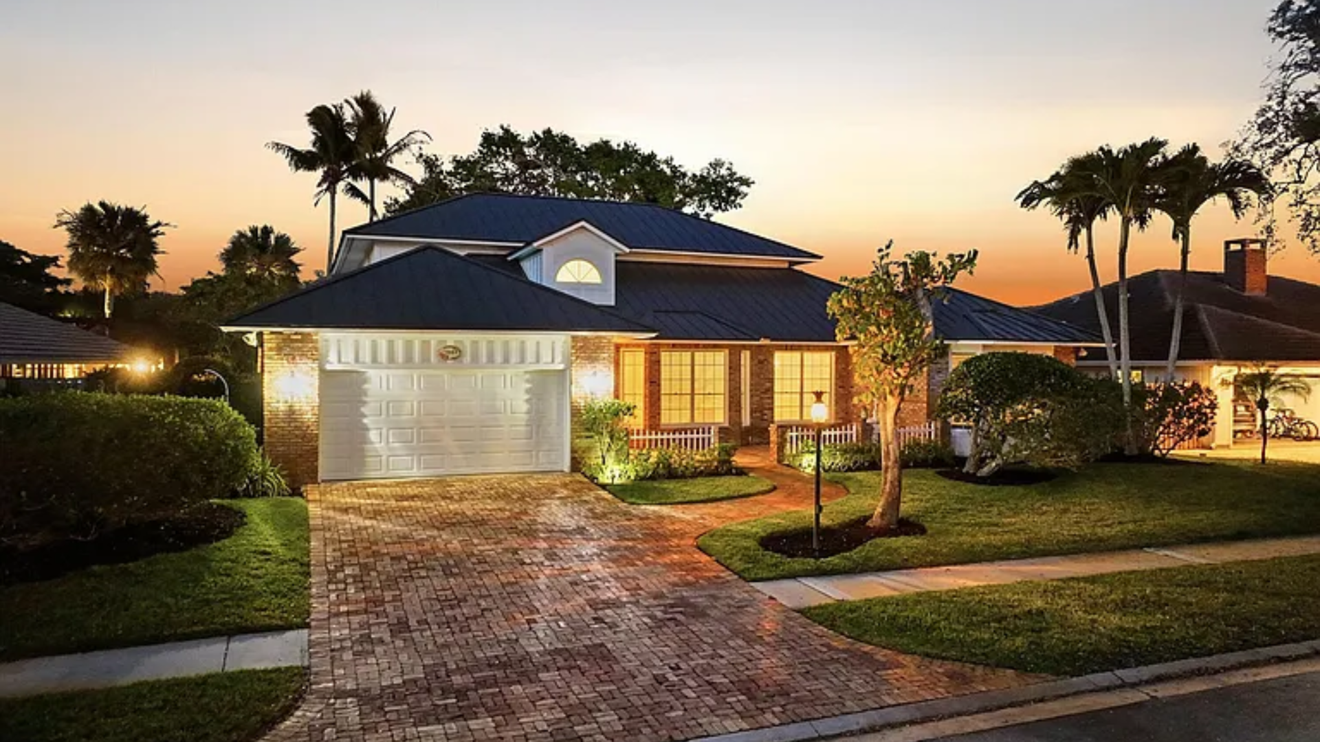 Suburban house at dusk with lit windows, driveway, and palm trees in front yard
