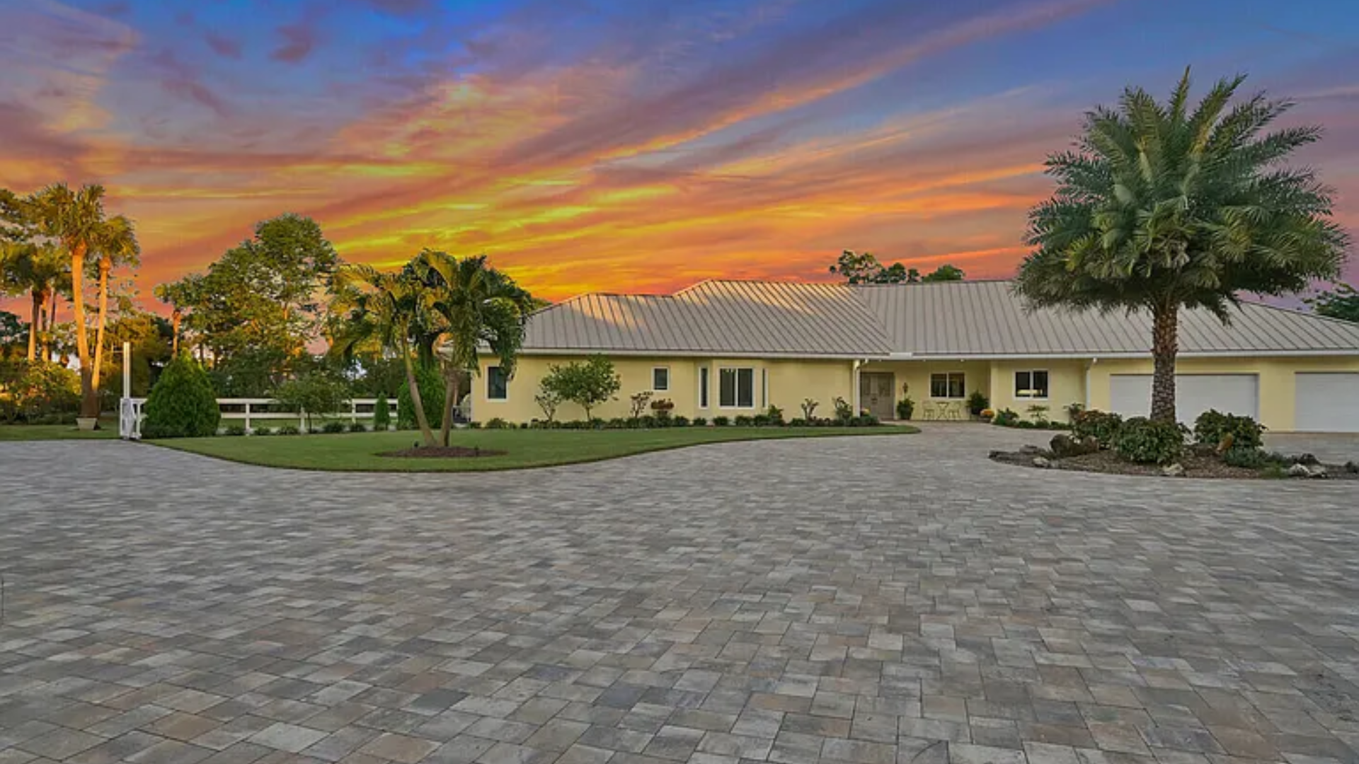 Sunset over a palm-lined driveway leading to a single-story home with a white fence and stone pavers.