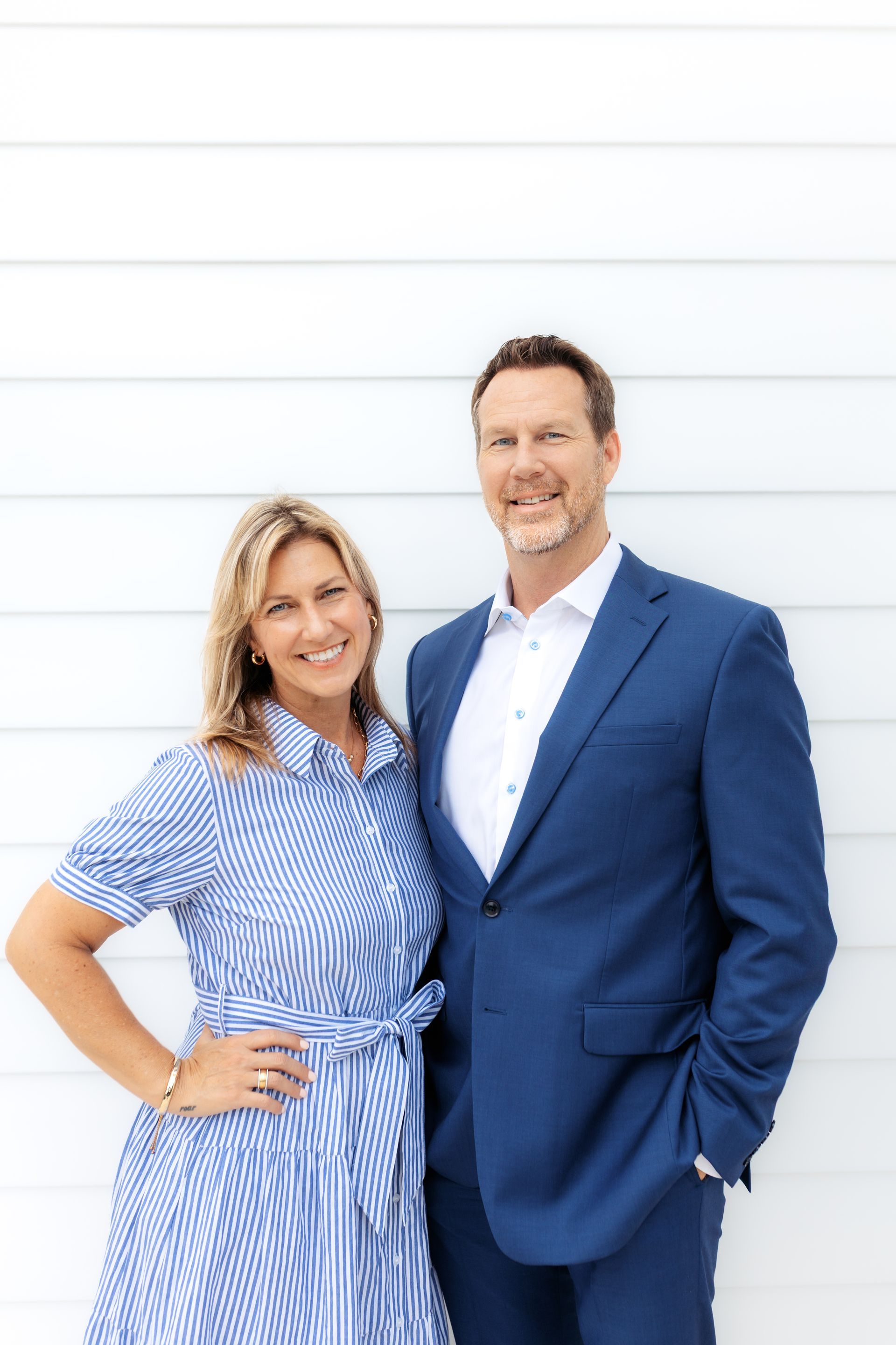 Two people standing together, smiling, against a white wall, wearing blue outfits.