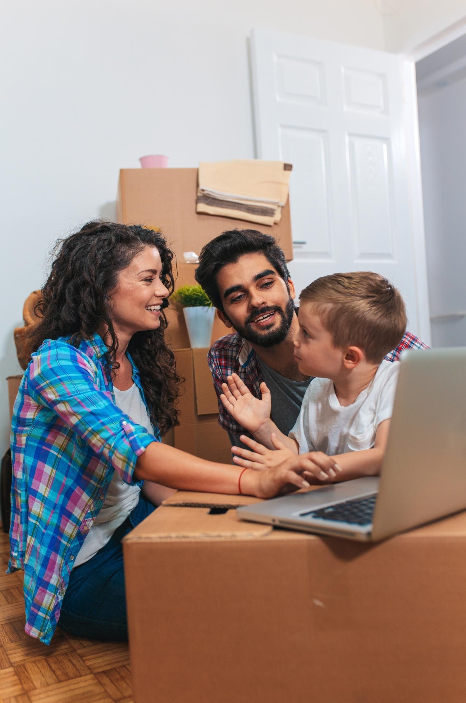 Family video chat with a smiling child and two adults beside a laptop in a moving box-filled room