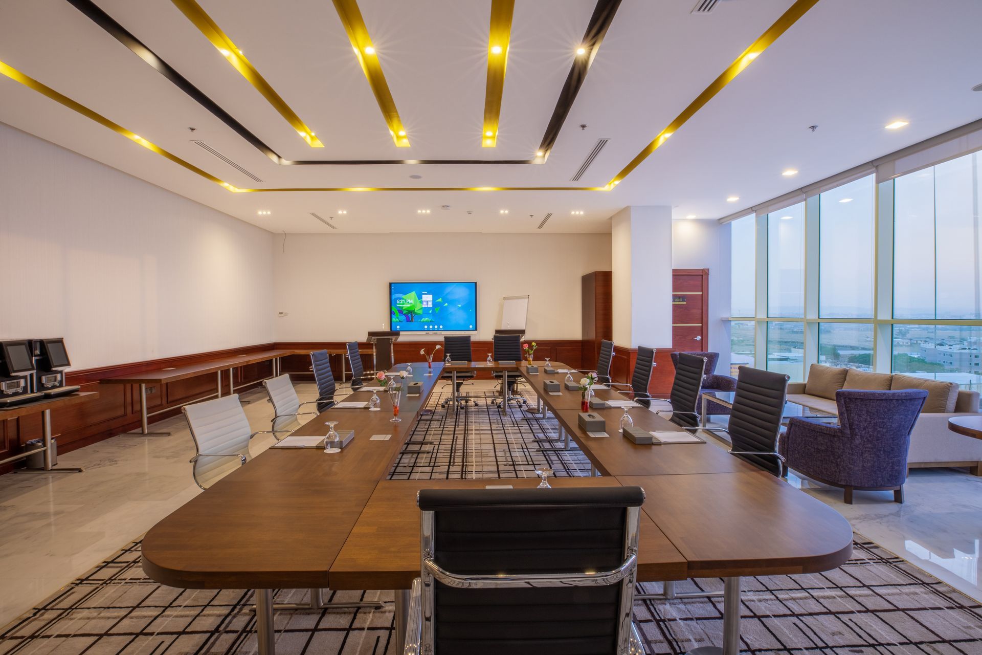 Modern conference room with a large oval table, chairs, wall screen, and floor-to-ceiling windows overlooking water