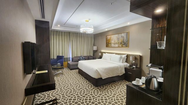 Modern hotel room with a king bed, patterned carpet, wall-mounted TV, and warm neutral decor