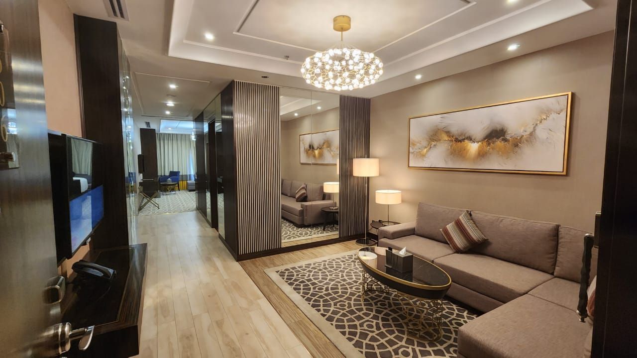 Modern living room with gray sectional sofa, patterned rug, chandelier, and wall-mounted TV alongside a hallway.