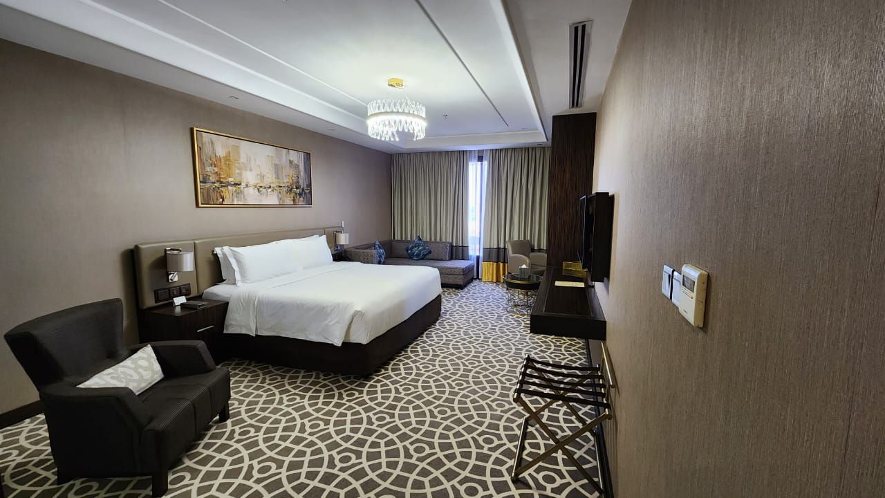 Modern hotel room with king bed, armchair, desk, patterned carpet, and window drapes