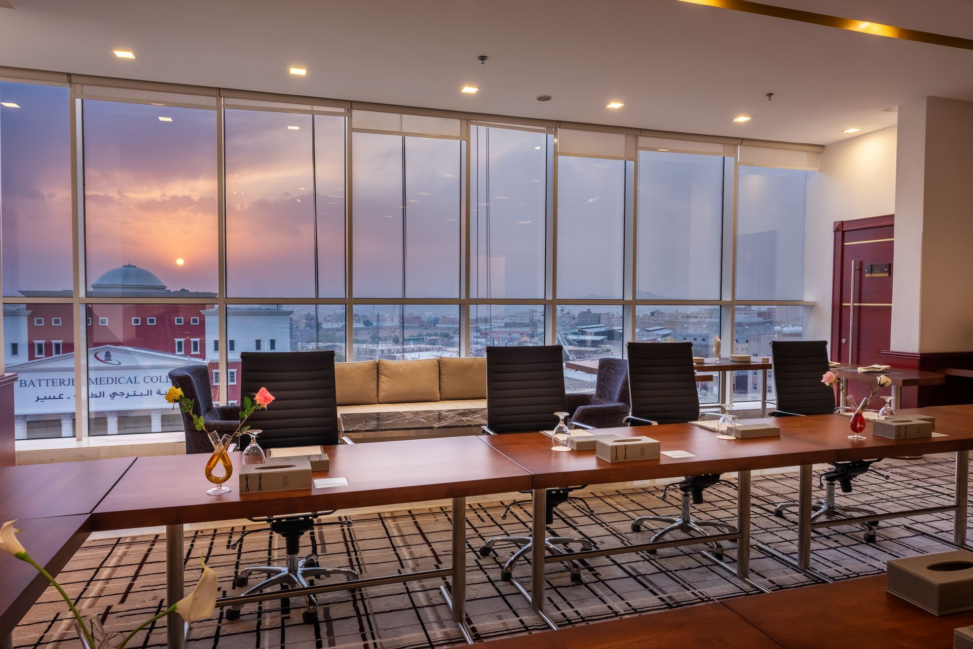 Modern conference room with long table, chairs, and floor-to-ceiling windows overlooking a city at sunset