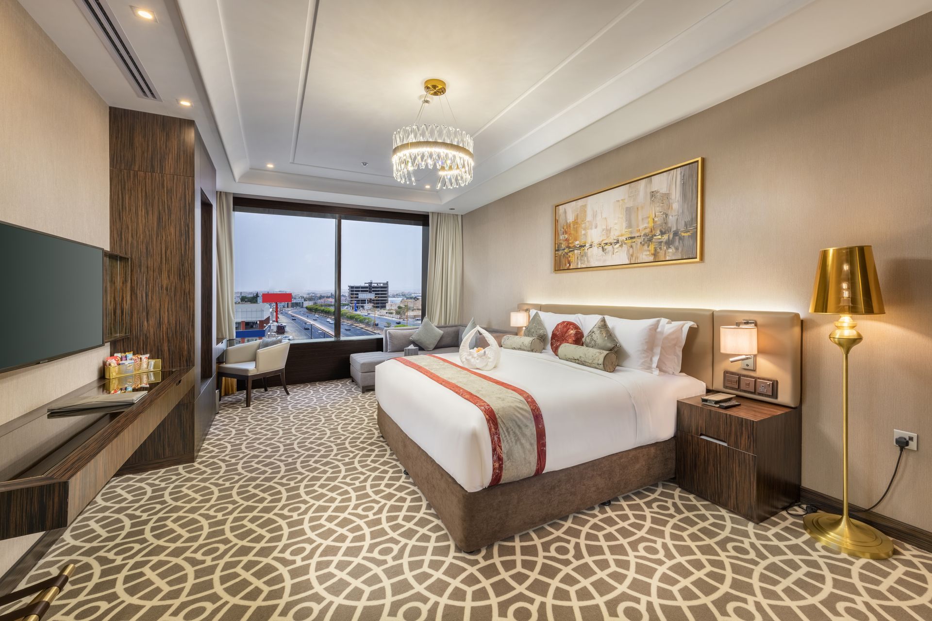 Modern hotel room with king bed, patterned carpet, chandelier, sofa, and city view through large window