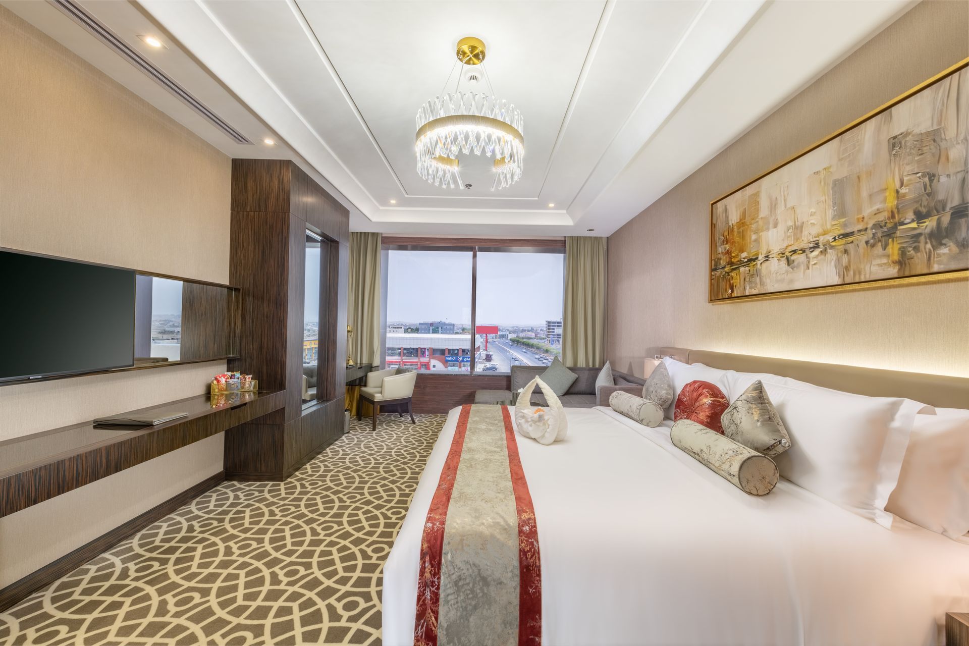 Hotel room with large bed, TV, window view, patterned carpet, and decorative chandelier.