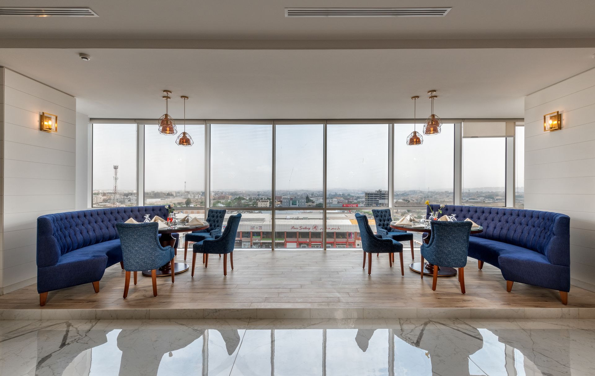 Modern lounge with blue velvet chairs, pendant lights, and large windows overlooking a city skyline.