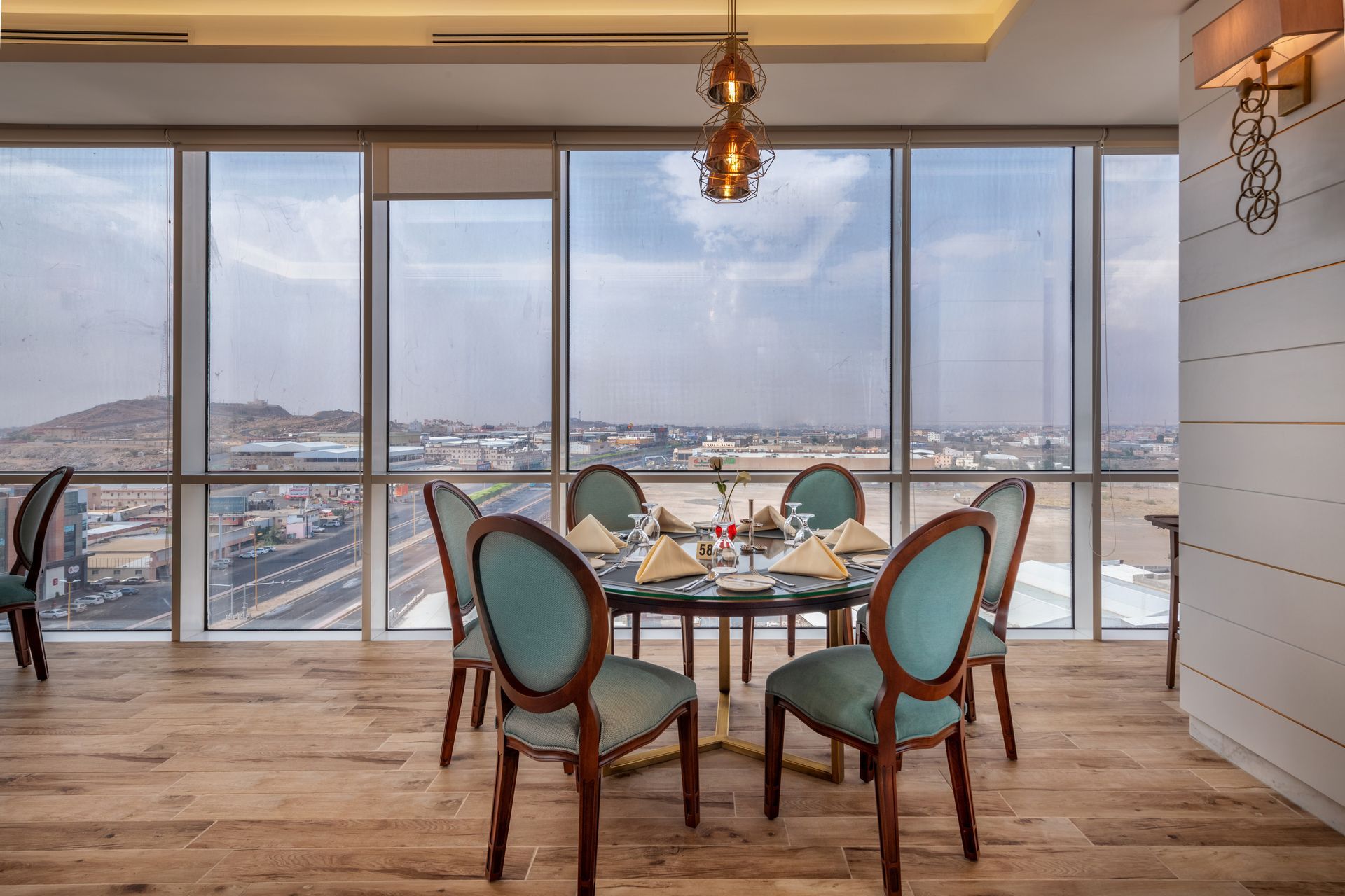 Modern dining room with glass table and teal chairs overlooking a city and hills through large windows