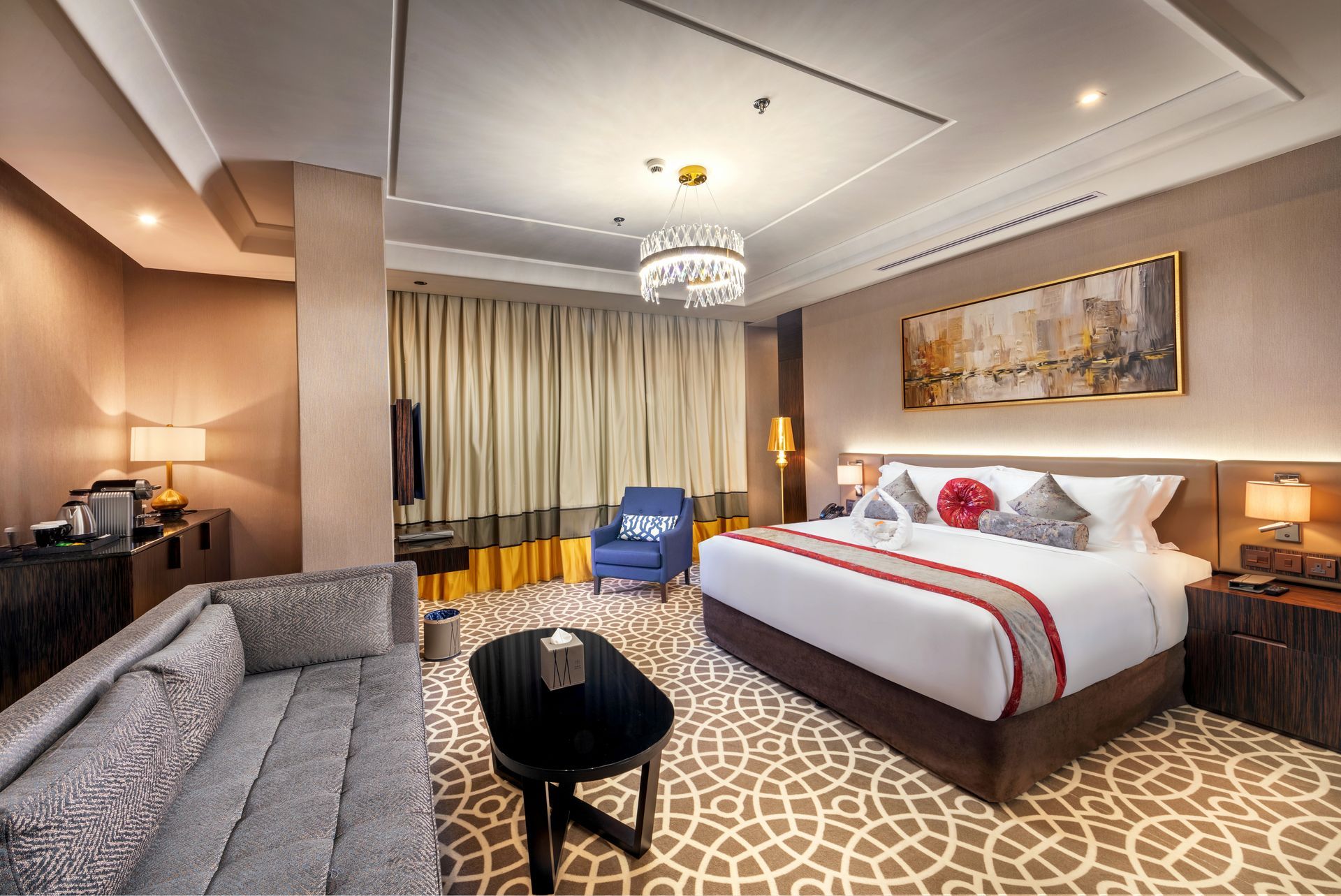 Hotel room interior with bed, couch, armchair, and coffee table. Neutral tones with patterned carpet.