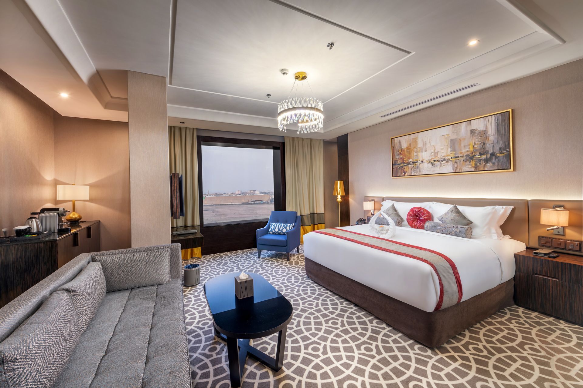 Modern hotel room with king bed, sofa, chair, patterned carpet, and city-view window