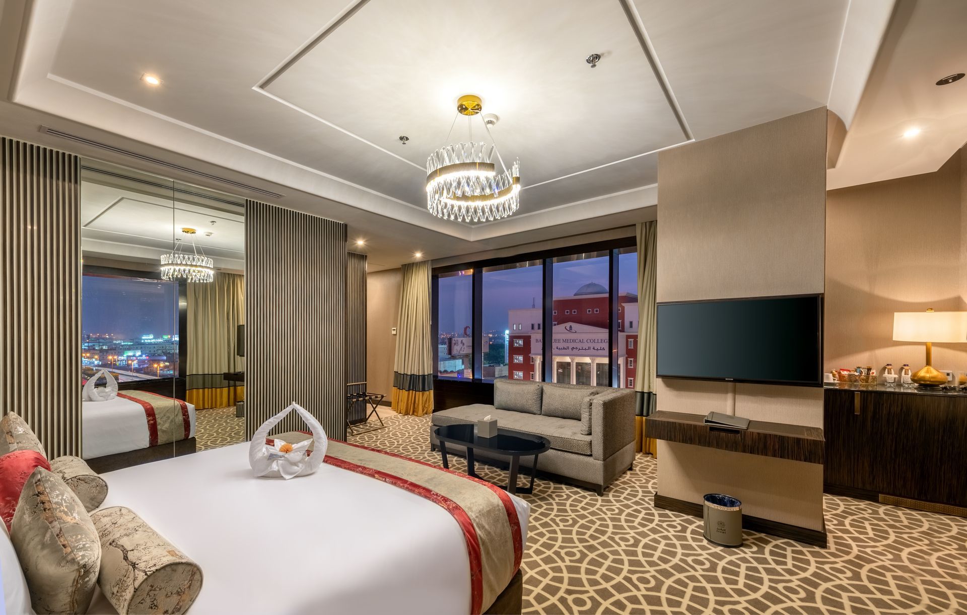 Modern hotel room with king bed, sofa, desk, chandelier, and city view windows