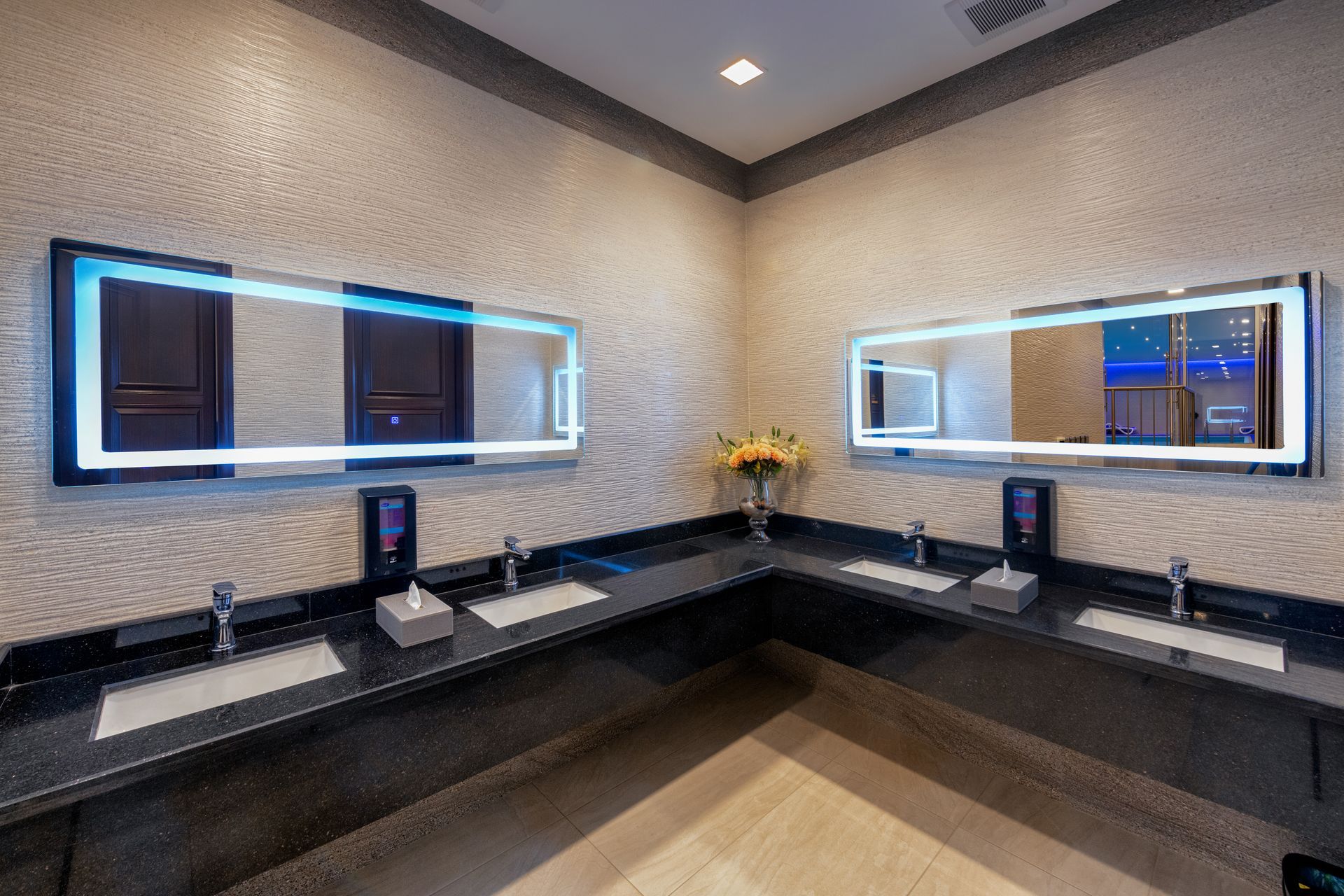 Corner bathroom with two sinks, black countertops, mirrors with blue lighting, and textured walls.