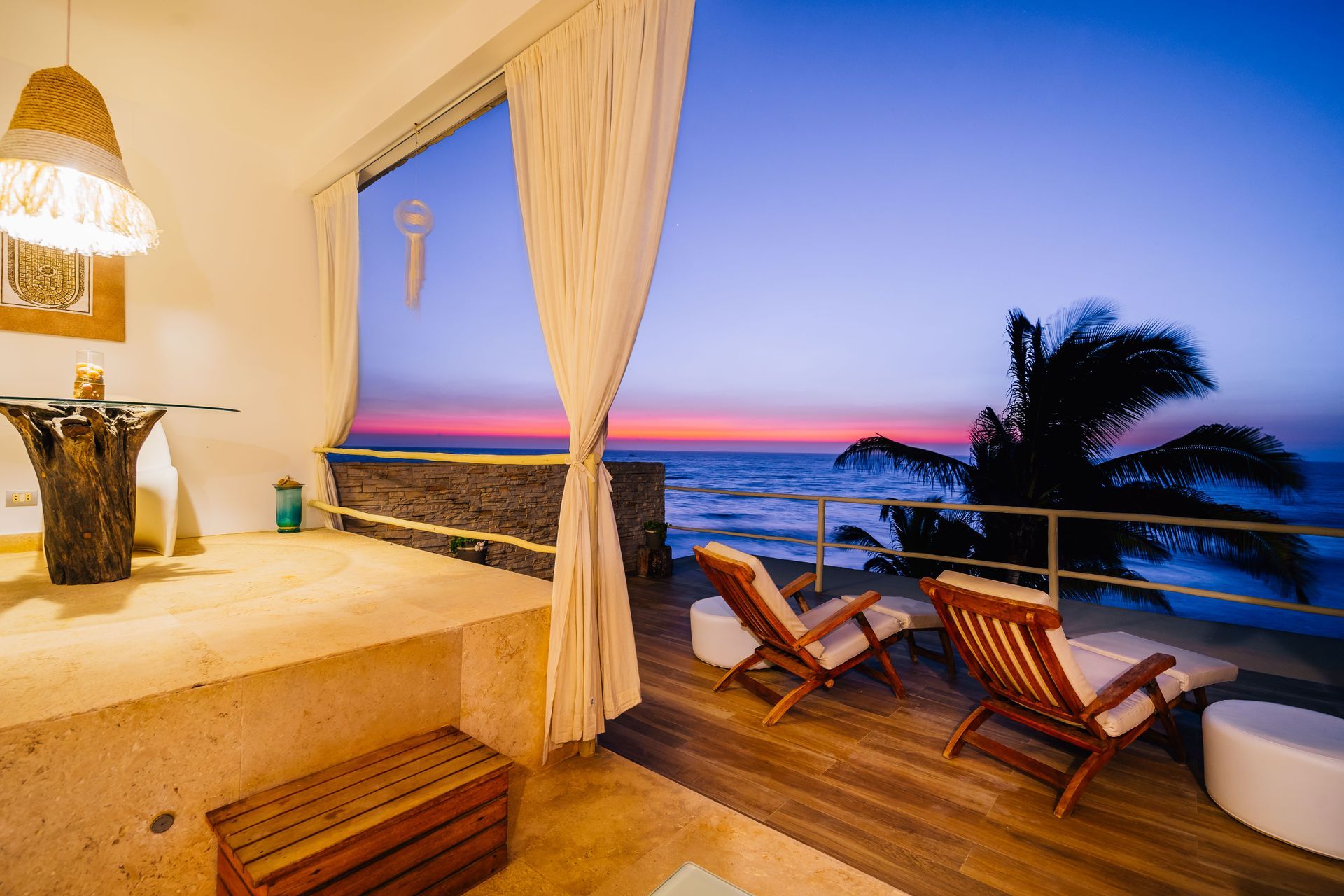 A balcony with chairs and a table overlooking the ocean at sunset.