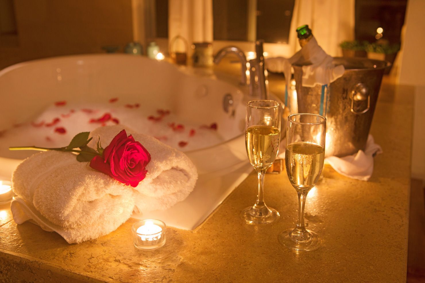 There is a bathtub with a rose in it and two glasses of champagne on the counter.