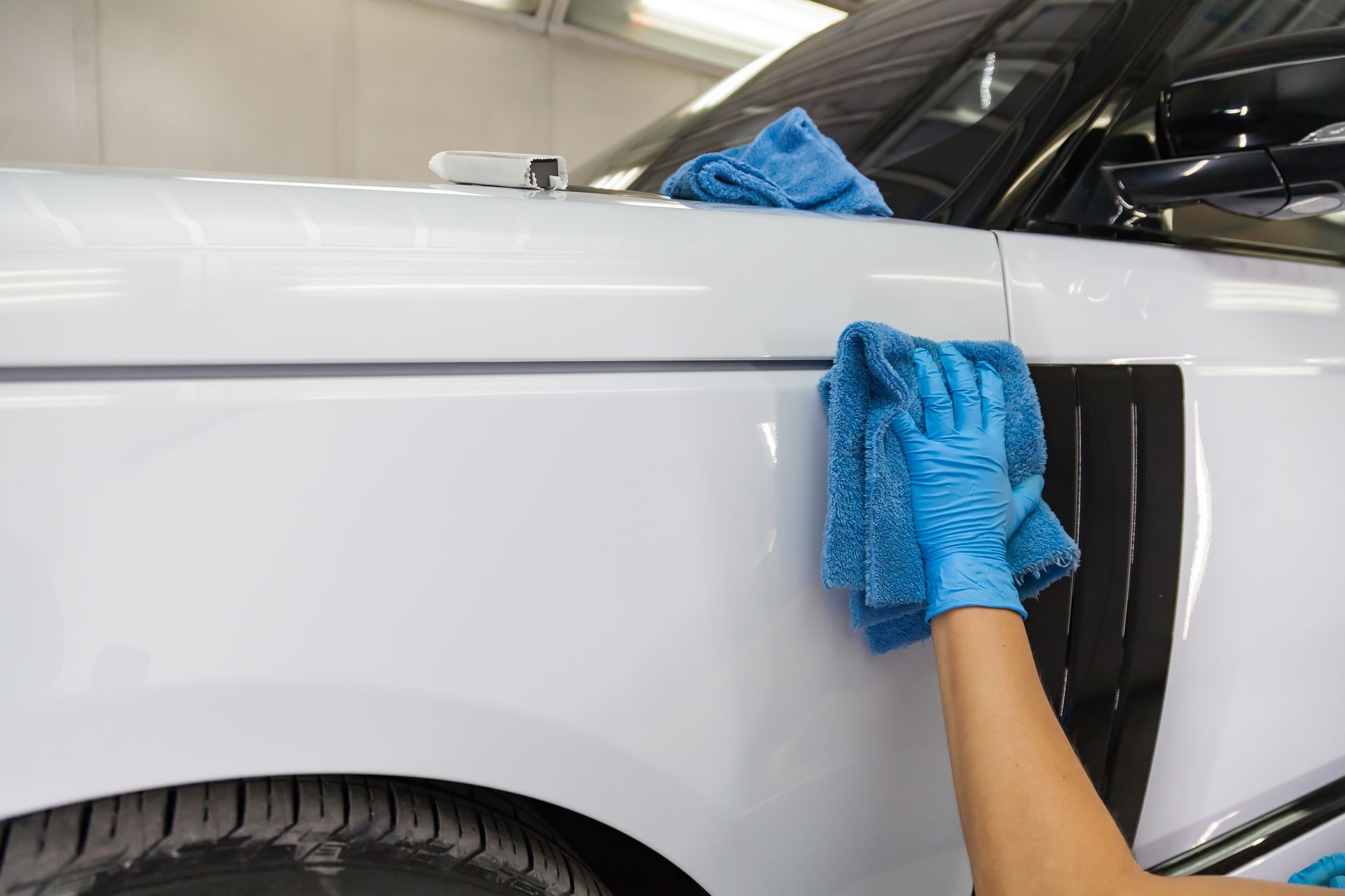 Person wiping a white car with a blue cloth in a garage, cleaning the front fender