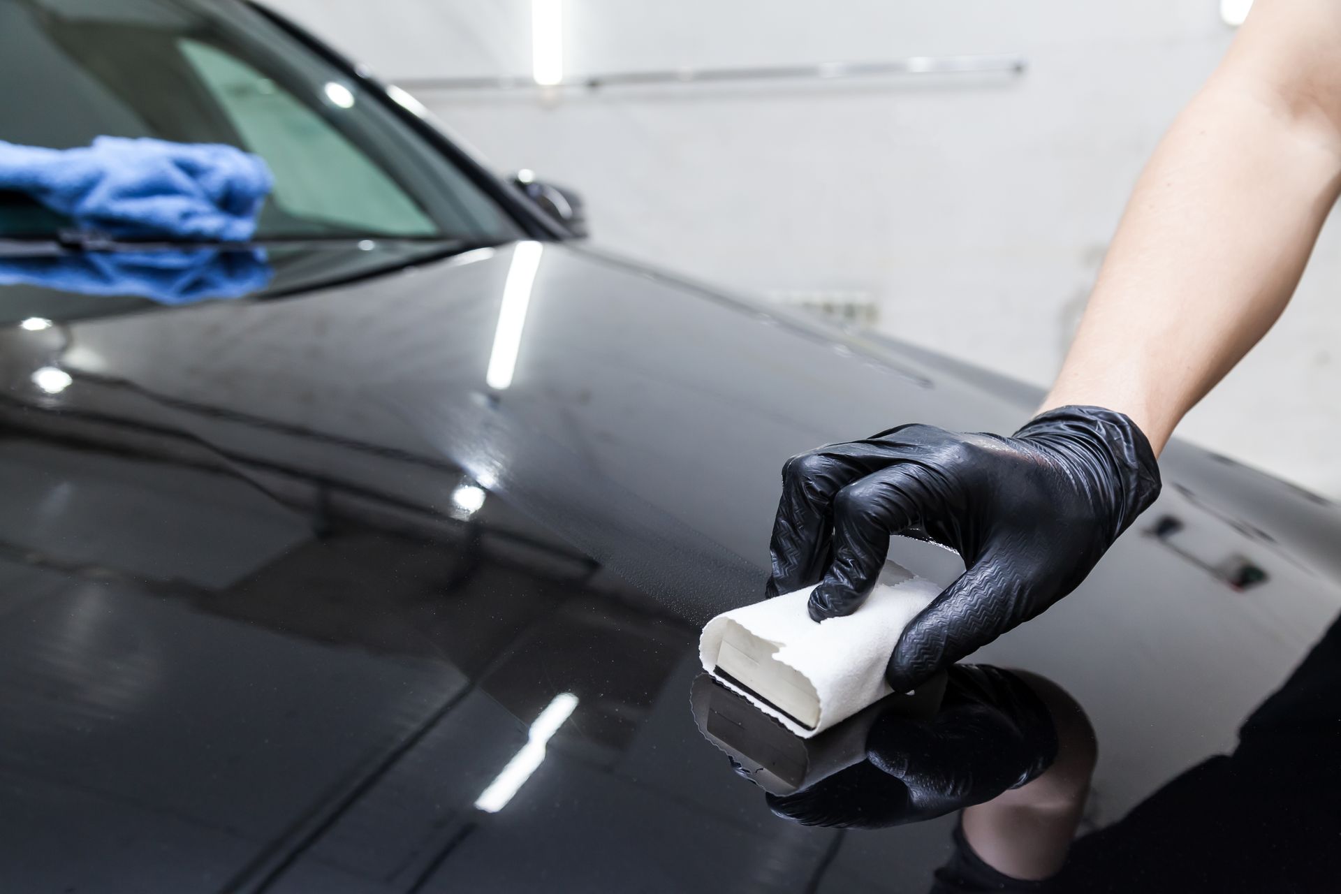 Black car hood being polished by a gloved hand with a white applicator in a garage