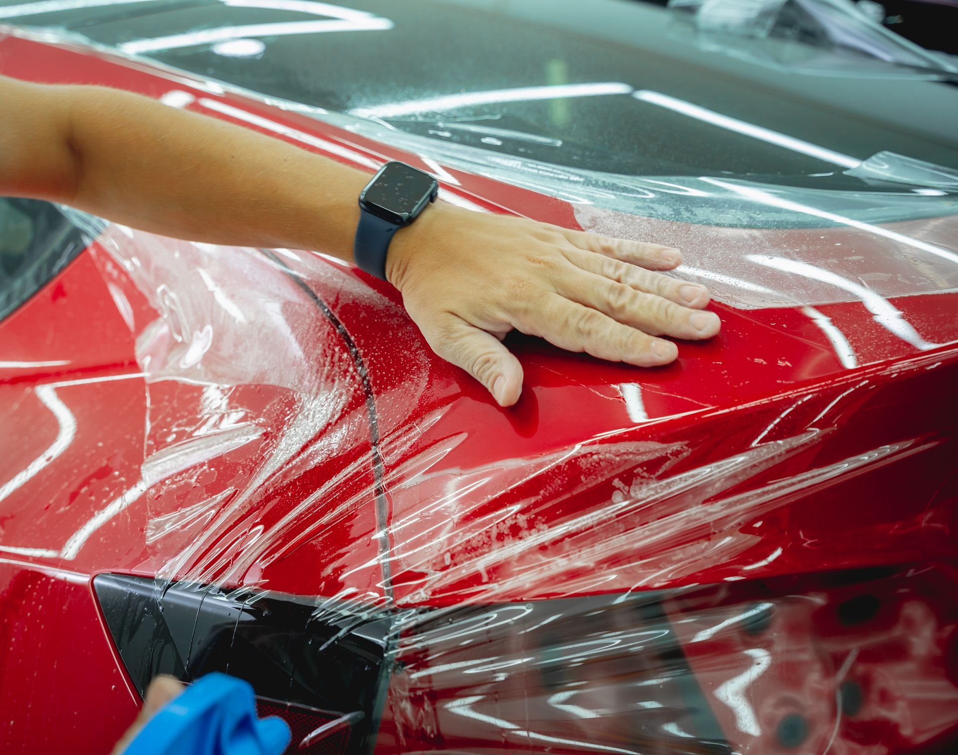 Hands applying protective film to a red car hood in a workshop