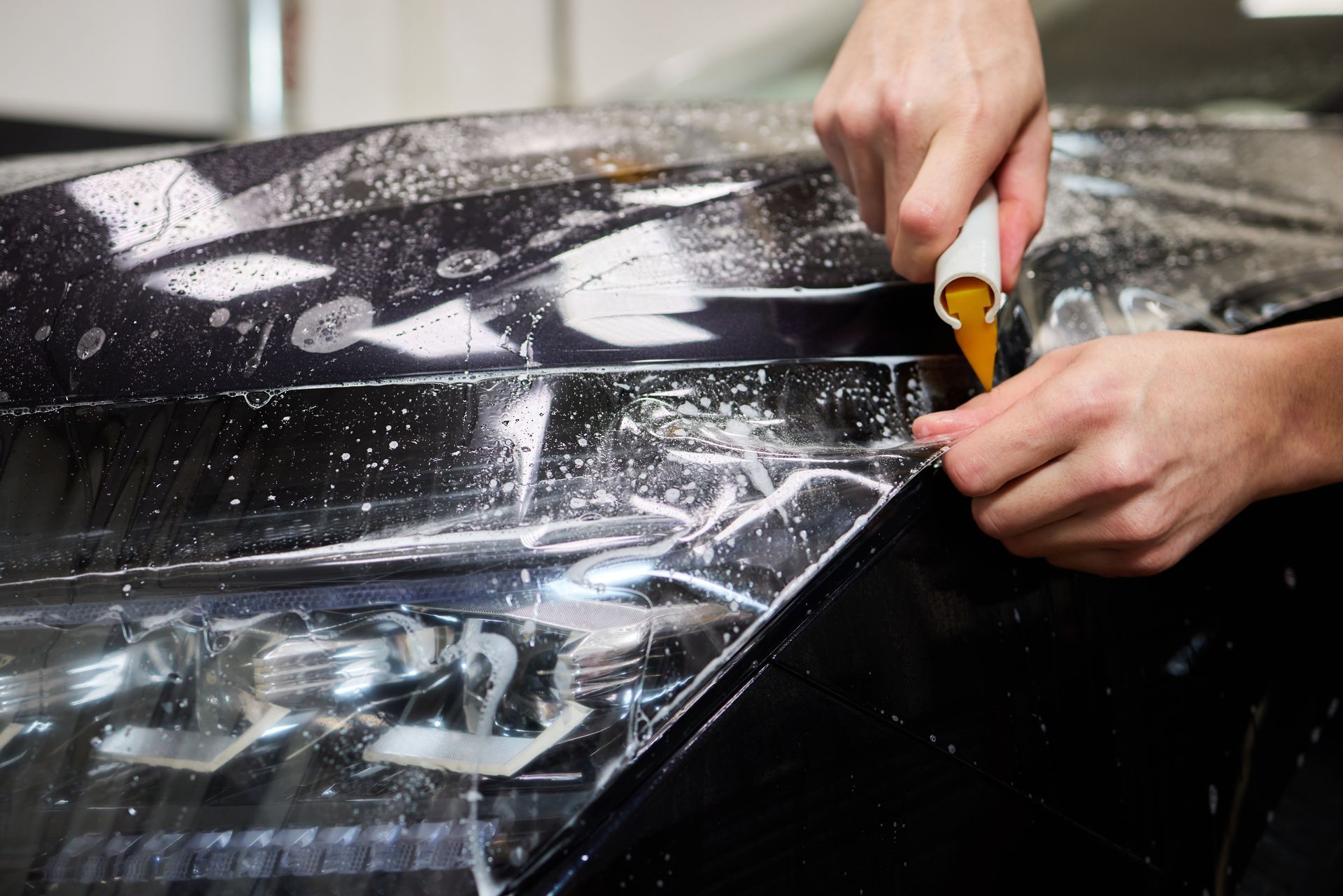 Hands using a tool to peel clear protective film from a black car hood.